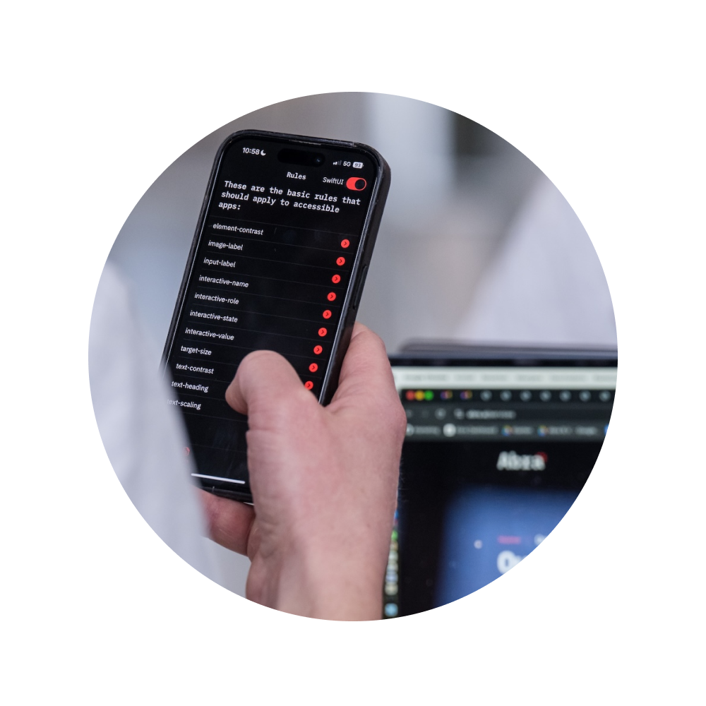 Hand holding a smartphone displaying a list of mobile app accessibility rules, with a laptop in the background.